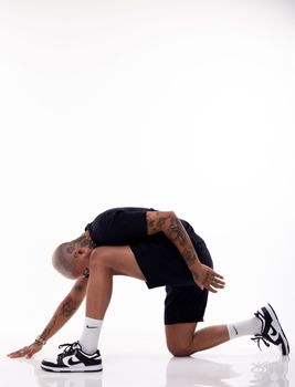 Side view of ethnic male with short hair and tattoos in sportswear standing on knee in white studio and looking down