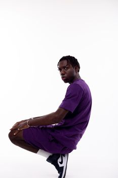 Side view of African American male with dreadlocks in sportswear squatting down in studio against white background and looking at camera
