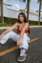 Charismatic young female with long dark hair in stylish clothes sitting on asphalt road on street in tropical city and looking at camera in daylight