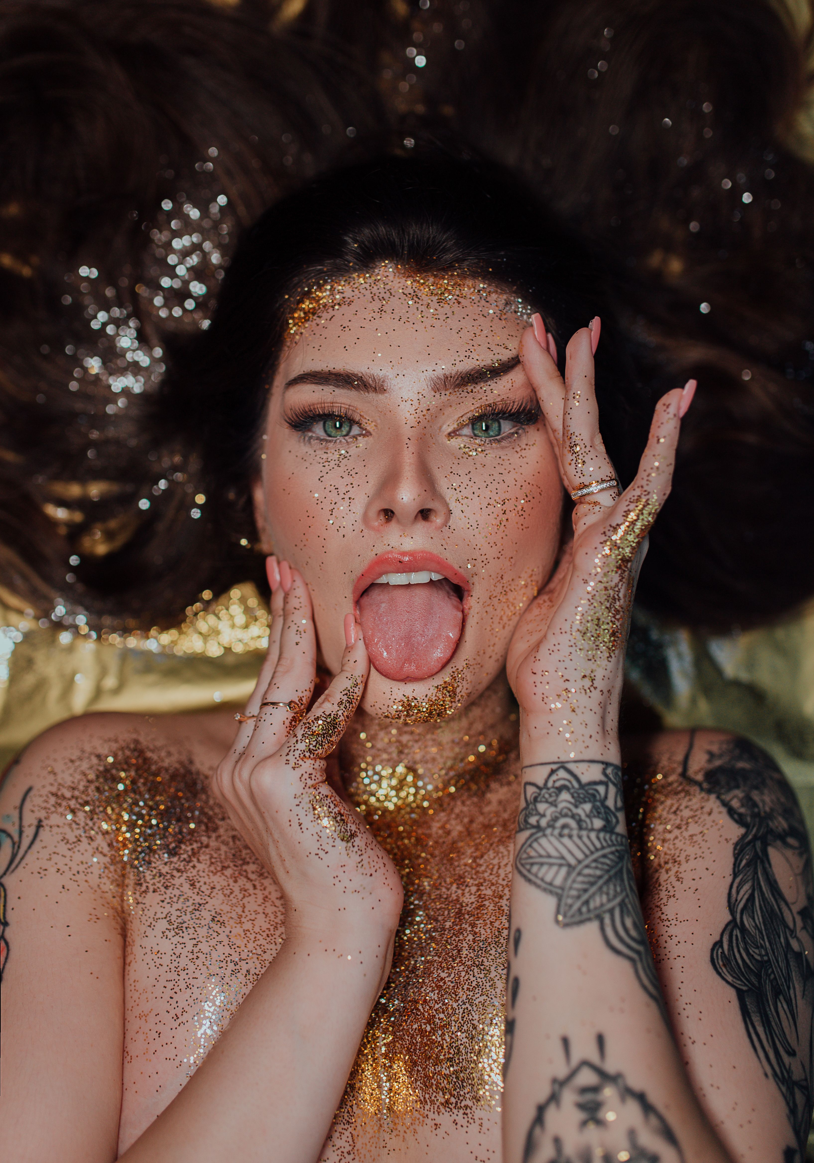 From above of topless young woman with long brown hair and tattoos covered with golden glitters lying with tongue out and touching face