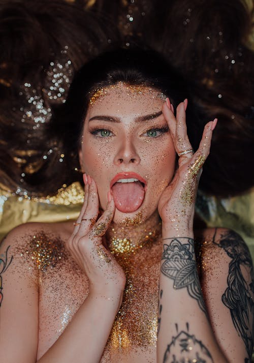 From above of topless young woman with long brown hair and tattoos covered with golden glitters lying with tongue out and touching face