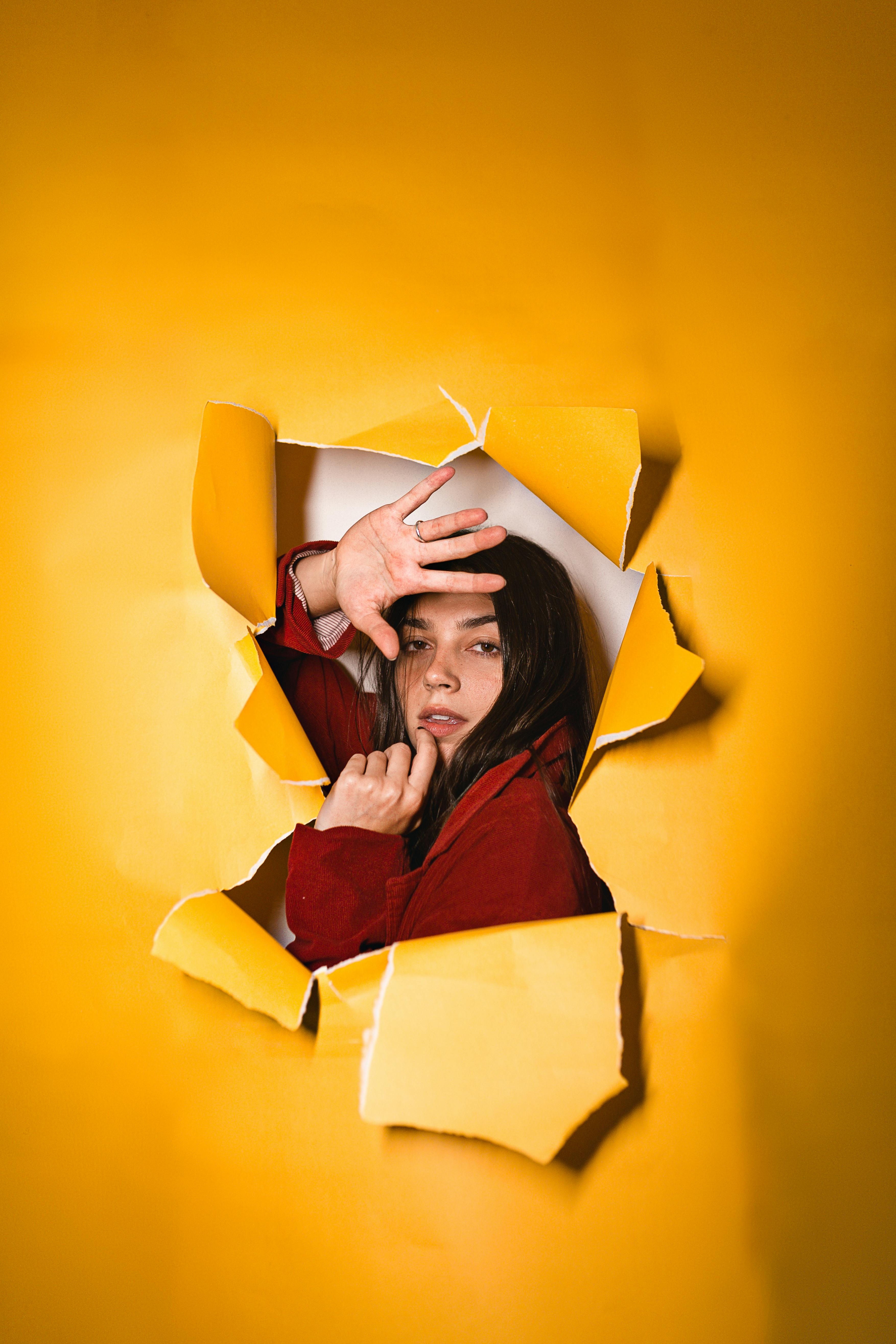 Young female with dark hair in red jacket raising arm and looking through hole in yellow paper background in studio