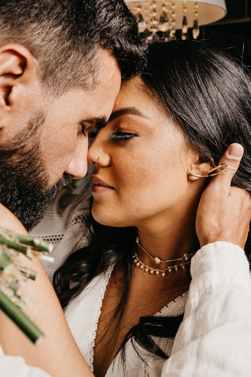 Crop adult bearded ethnic groom embracing feminine bride with piercing and closed eyes on wedding day