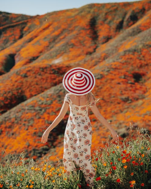 Woman In Floral Dress With Red and White Sun Hat Standing Beside Blooming Flowers 