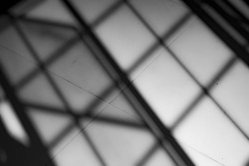 Black and white from below of white tiled ceiling with shadow of geometric metal grid in light room of building