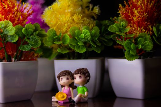 Cute couple of boy and girl figurines placed on dark table near white flowerpots with flowers and green leaves in room