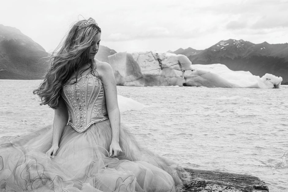 Black and white portrait of a woman in a gown by icy shore, with mountains.