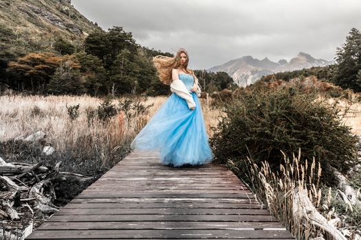 A woman in a flowing blue gown poses on a boardwalk surrounded by nature, exuding elegance.