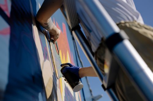 Free From below of crop unrecognizable person in casual clothes and gloves using spray can while painting graffiti wall standing on ladder Stock Photo