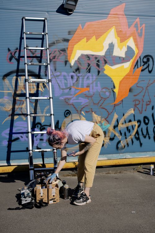 Free Side view of young tattooed female millennial in stylish clothes choosing paint cans while painting graffiti wall on city street on sunny day Stock Photo