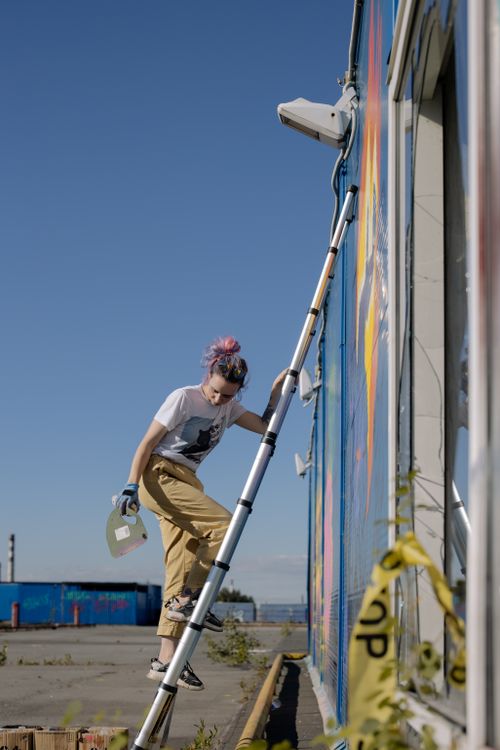 Free A young woman paints a large mural on a building wall using a ladder on a sunny day. Stock Photo
