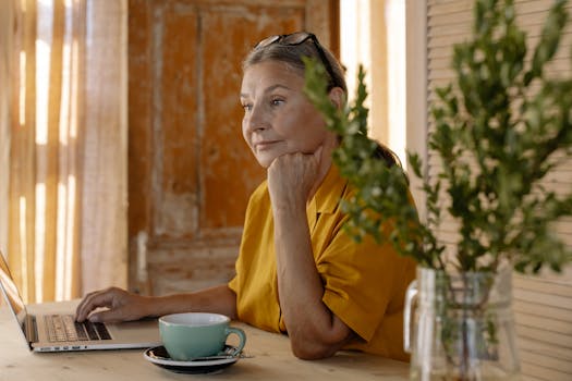 Elderly woman sitting at a table using a laptop, relaxing in a cozy home setting.
