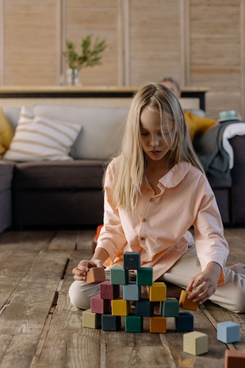 Free A young girl in pink pajamas plays with colorful stacking blocks on a wooden floor in a cozy living room. Stock Photo