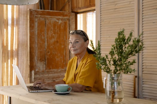 Senior woman in a yellow shirt working on a laptop at home, enjoying a coffee.