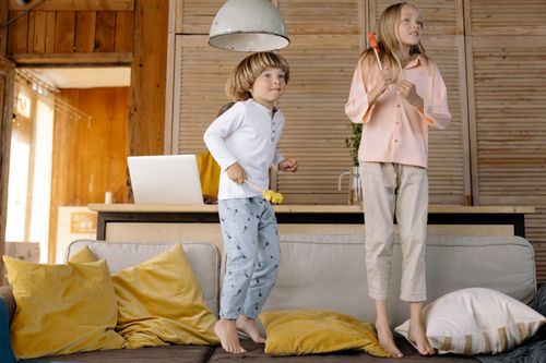 Free Two kids having fun playing and jumping on a cozy sofa indoors with pillows scattered around. Stock Photo