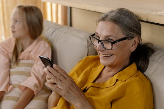 Senior woman in glasses smiling while using smartphone indoors, sitting with a child.