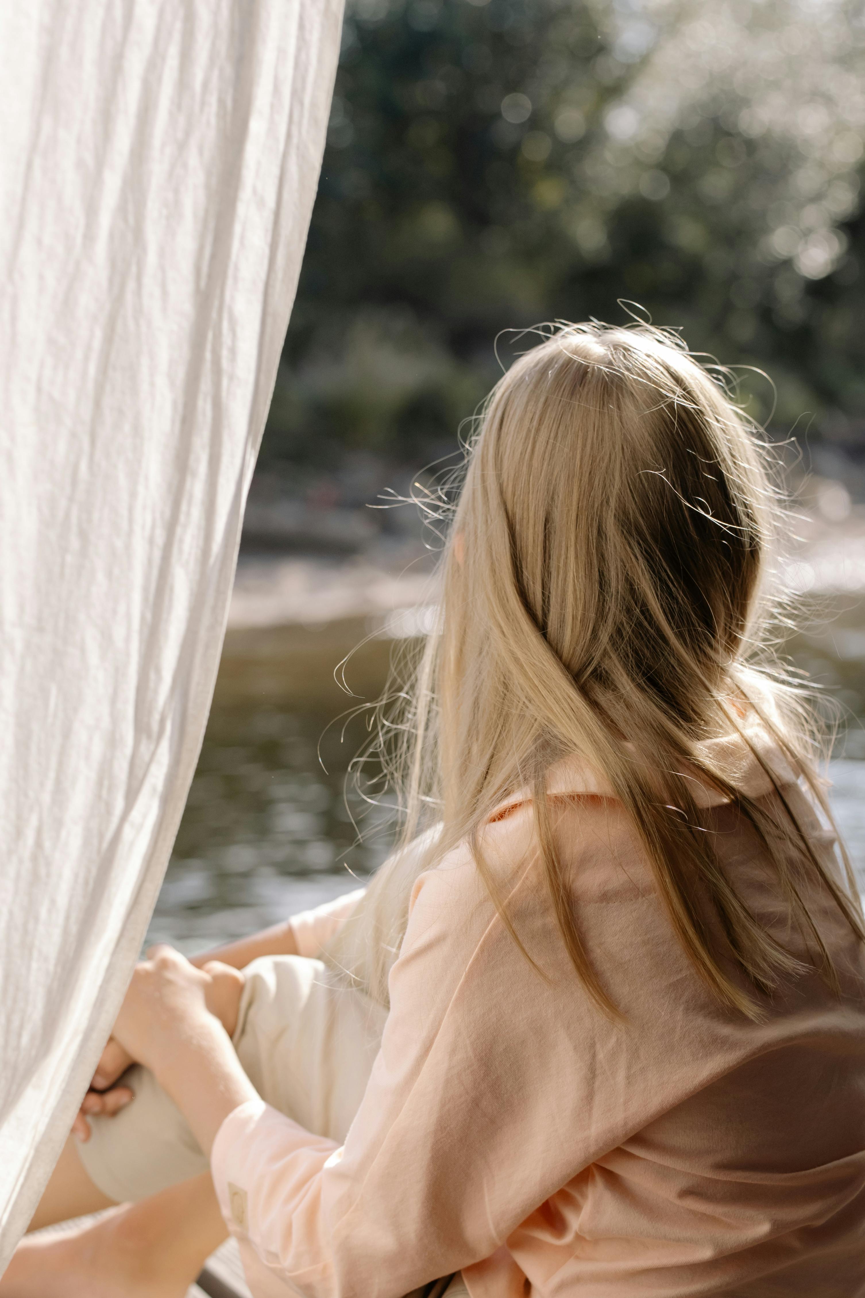A blonde child sits by a window, gazing outside in a serene indoor setting.