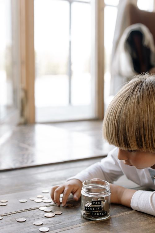 Free A young boy counting coins and placing them into a jar labeled 'for barbie castle' indoors on wooden flooring. Stock Photo