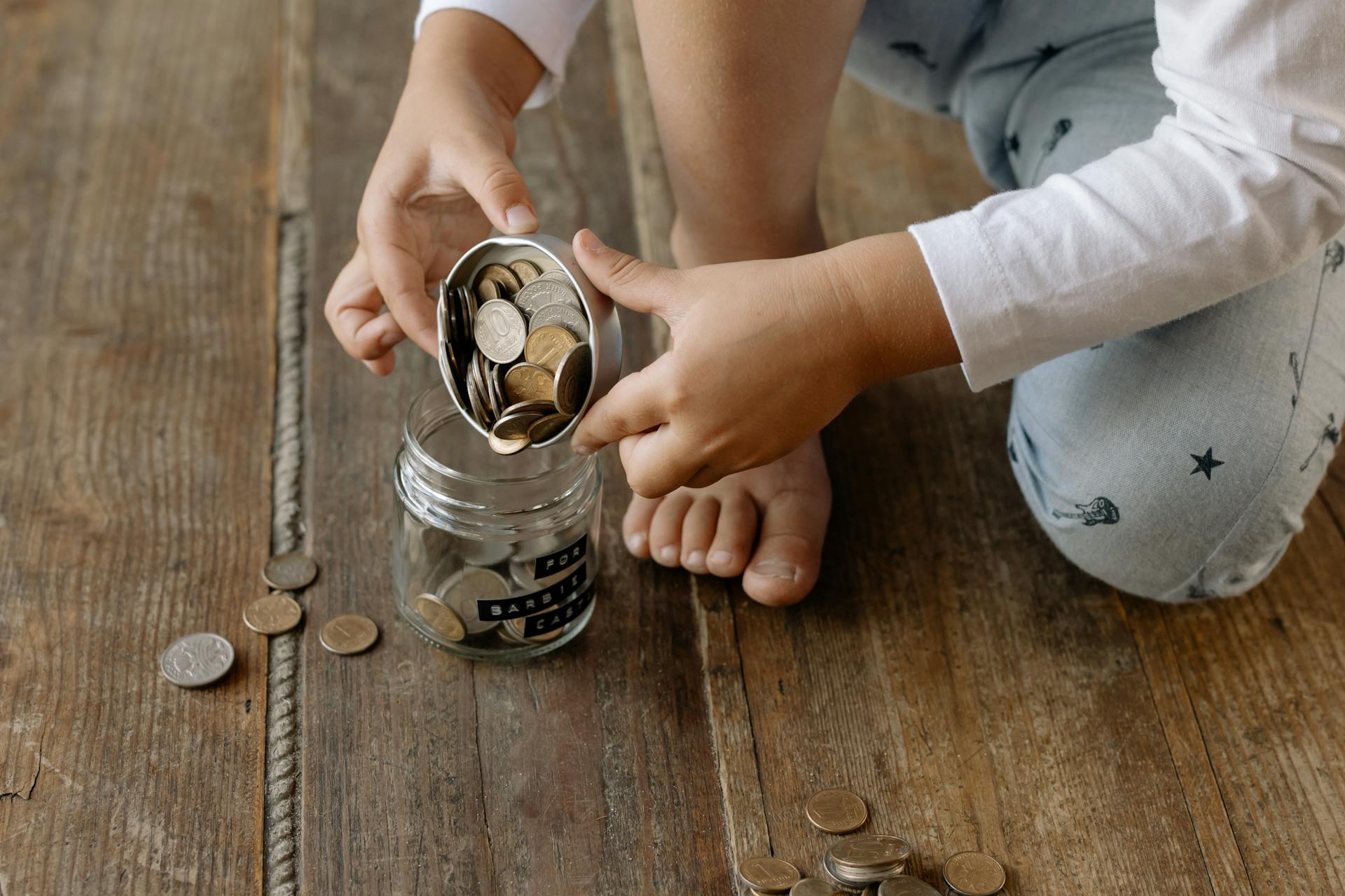 A child adds coins into a glass jar labeled for savings on a wooden floor.