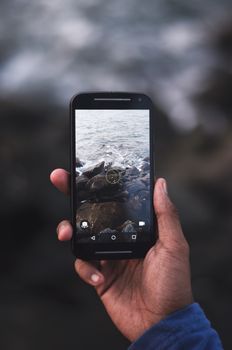 A hand holds a smartphone capturing the rocky seaside landscape in focus.