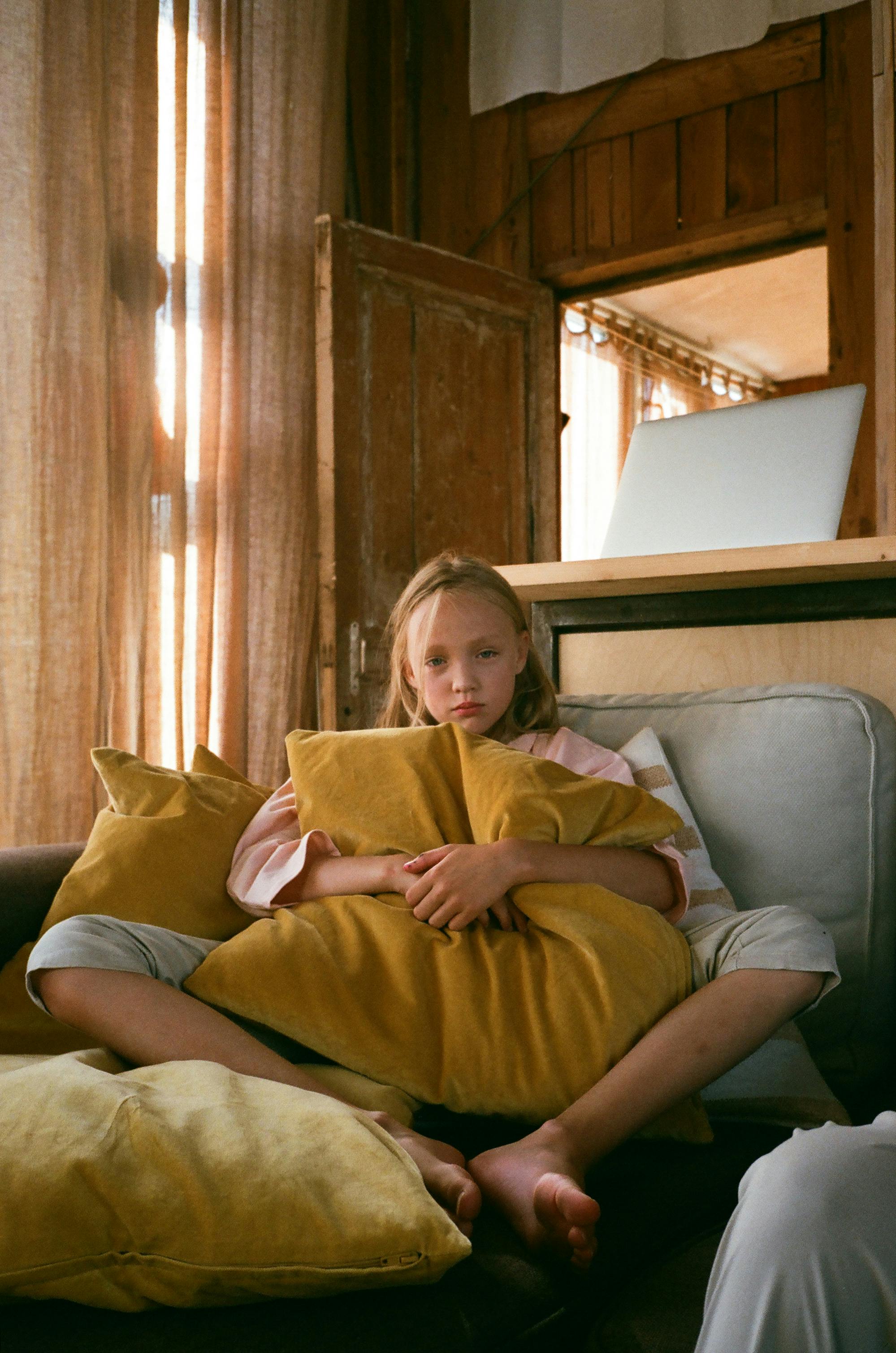 Young girl sitting comfortably on a couch, hugging a cushion indoors.