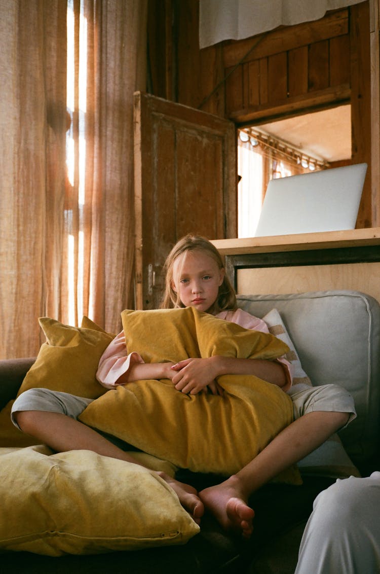 A Young Girl Sitting On The Couch While Hugging A Pillow