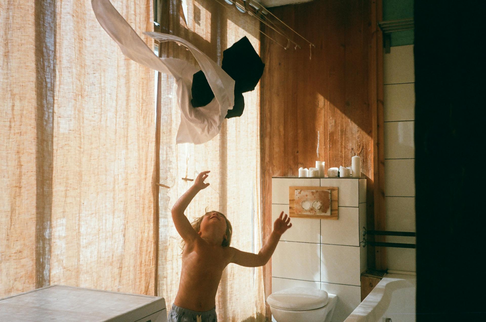 Child throws clothes in a sunlit bathroom, capturing carefree play and natural light.