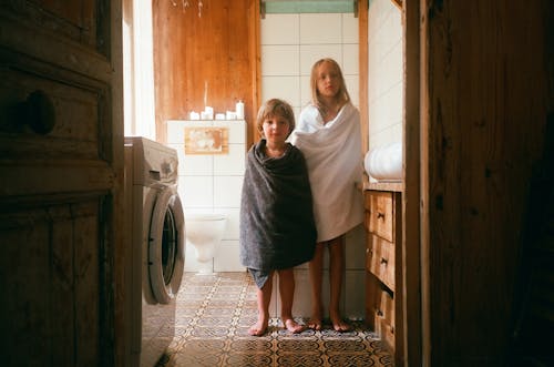 Photo of a Boy and a Girl with Bath Towels Standing in a Bathroom