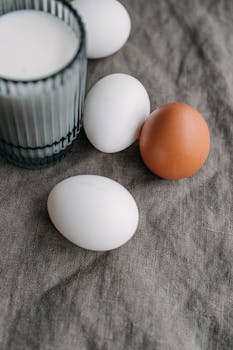 A close-up shot of brown and white eggs beside a glass of milk on fabric.