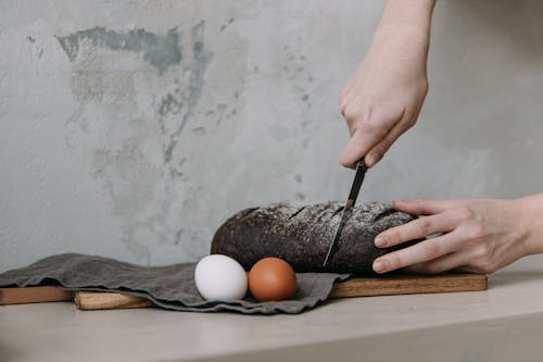 Photo of a Person's Hands Slicing a Black Loaf of Bread
