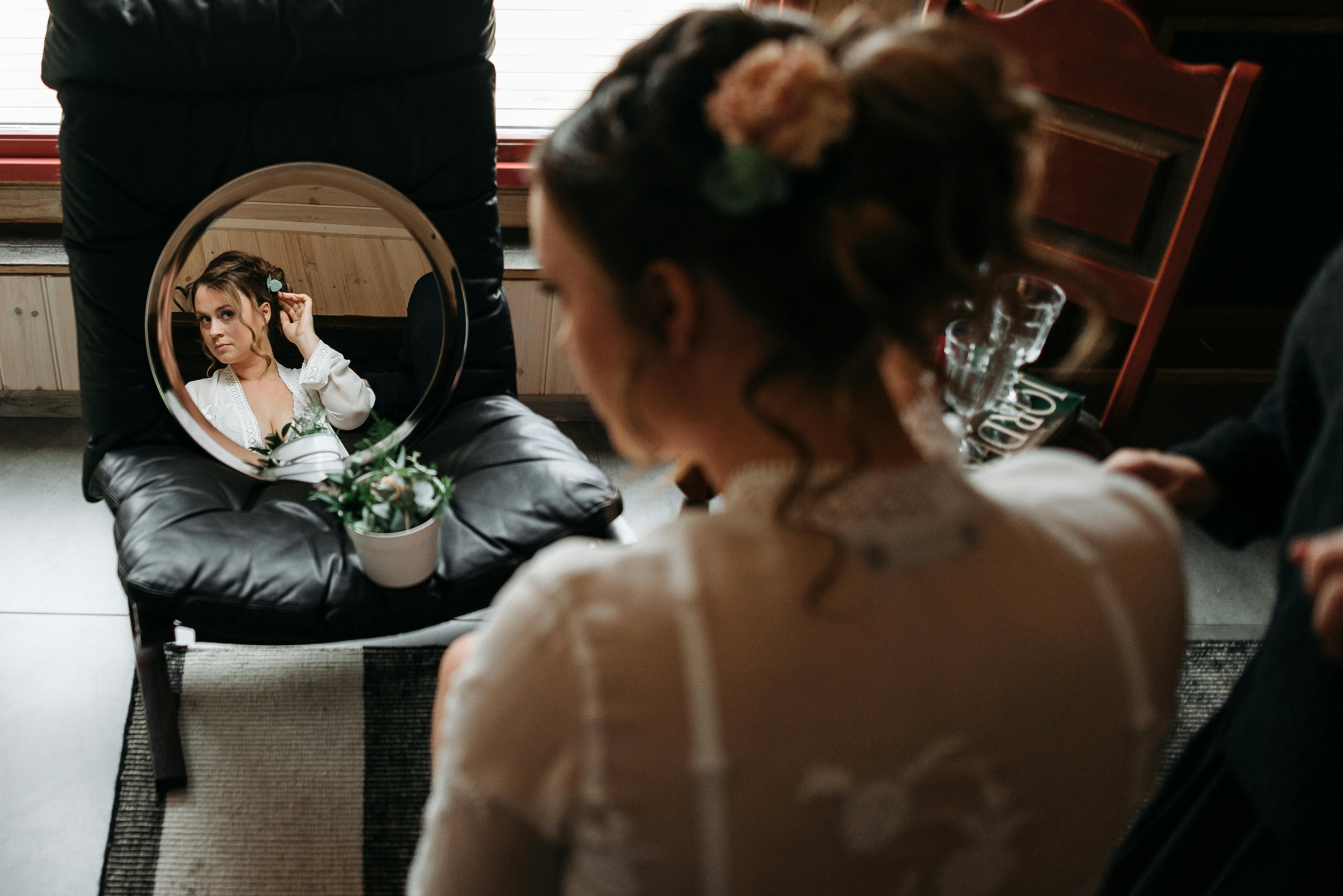 A bride adjusts her hair while looking into a round mirror indoors.