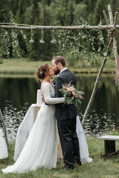 Bride and groom share a tender kiss during an outdoor wedding ceremony by the lakeside.