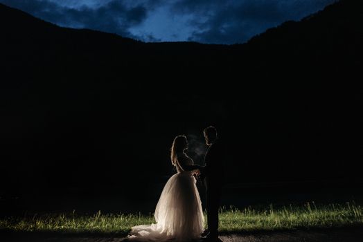 A romantic silhouette of a bride and groom holding hands at dusk in a mountainous setting.