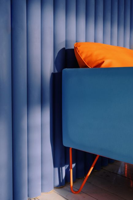 Stylish blue sofa with a vibrant orange pillow against a vertical blue curtain backdrop.