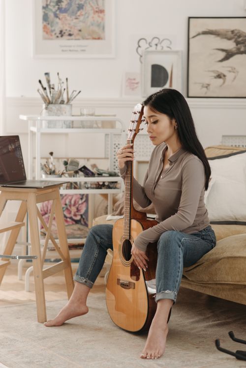 Free Asian woman seated in a cozy studio holding an acoustic guitar, surrounded by artistic elements. Stock Photo