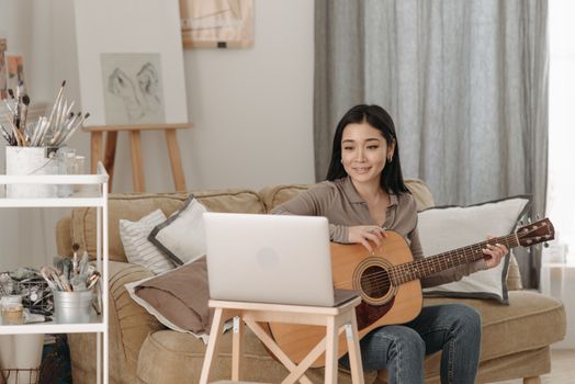 A woman playing guitar and learning through an online lesson at home using a laptop.