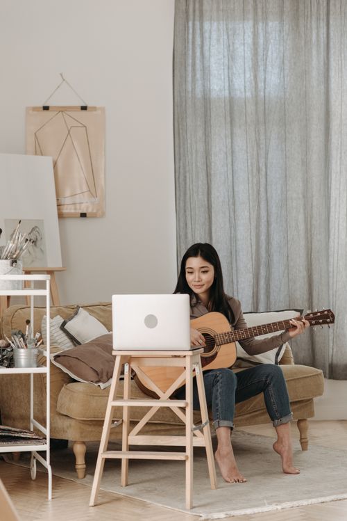 Free A woman learns guitar at home via online lessons using a laptop, sitting comfortably on a couch. Stock Photo