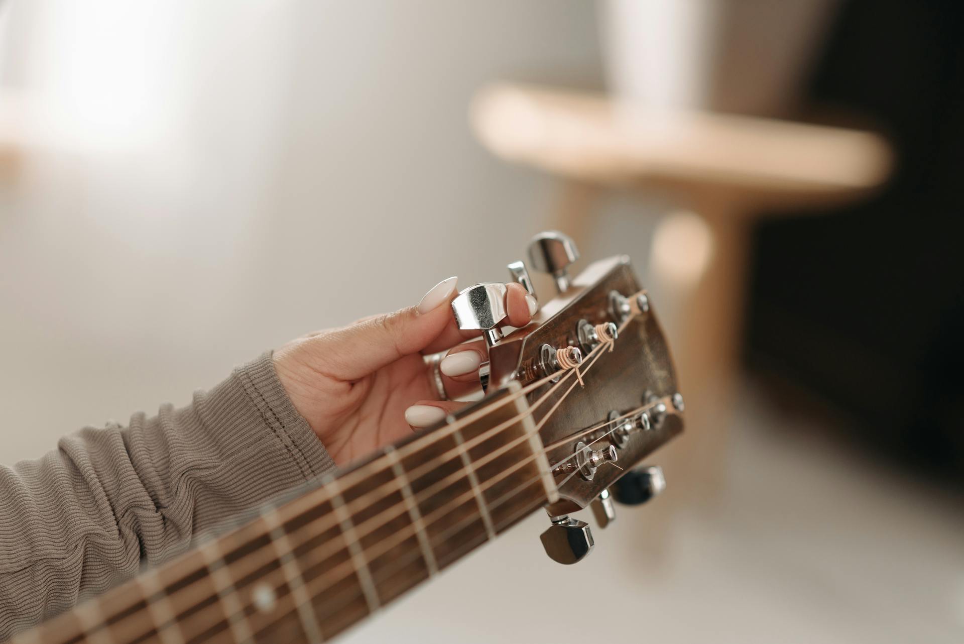 A detailed view of a person tuning an acoustic guitar indoors, focusing on the hand and guitar head.