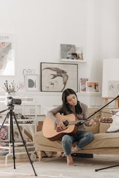 Asian woman playing guitar in a cozy room setup with a camera on a tripod, capturing her practice session.