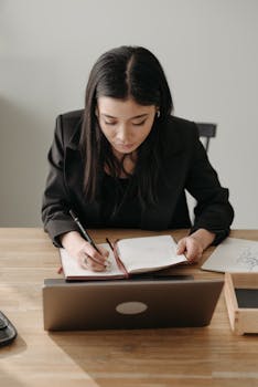 A woman in a black blazer taking notes at a desk with a laptop, emphasizing remote work.