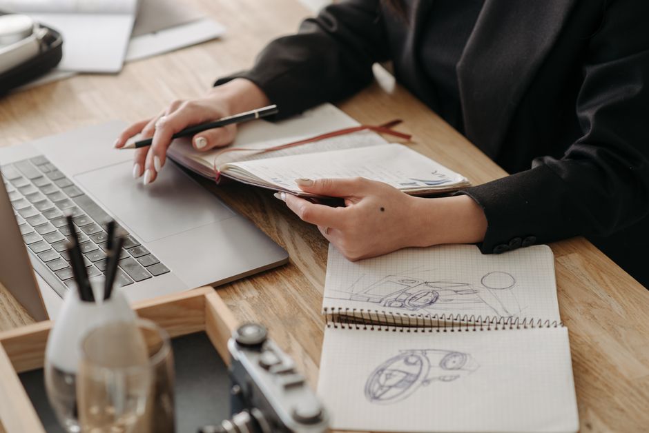 Desk with a laptop, sketches, and hands holding a notebook, embodying creative work.