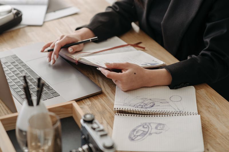 Desk with a laptop, sketches, and hands holding a notebook, embodying creative work.