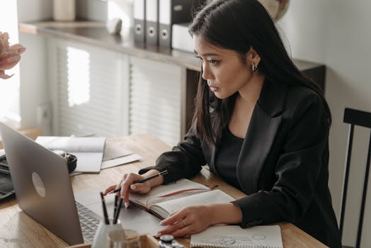 Young woman in blazer taking notes during an online lecture at home workspace.