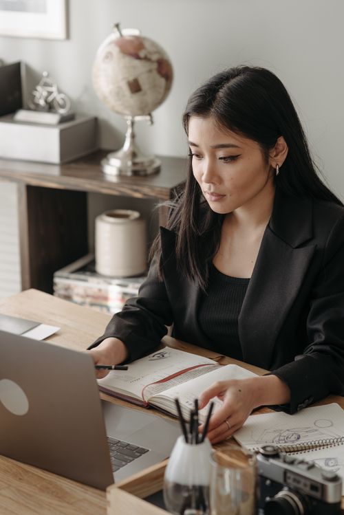Free Asian woman in office attire focused on work at a desk with a laptop and notebook. Stock Photo