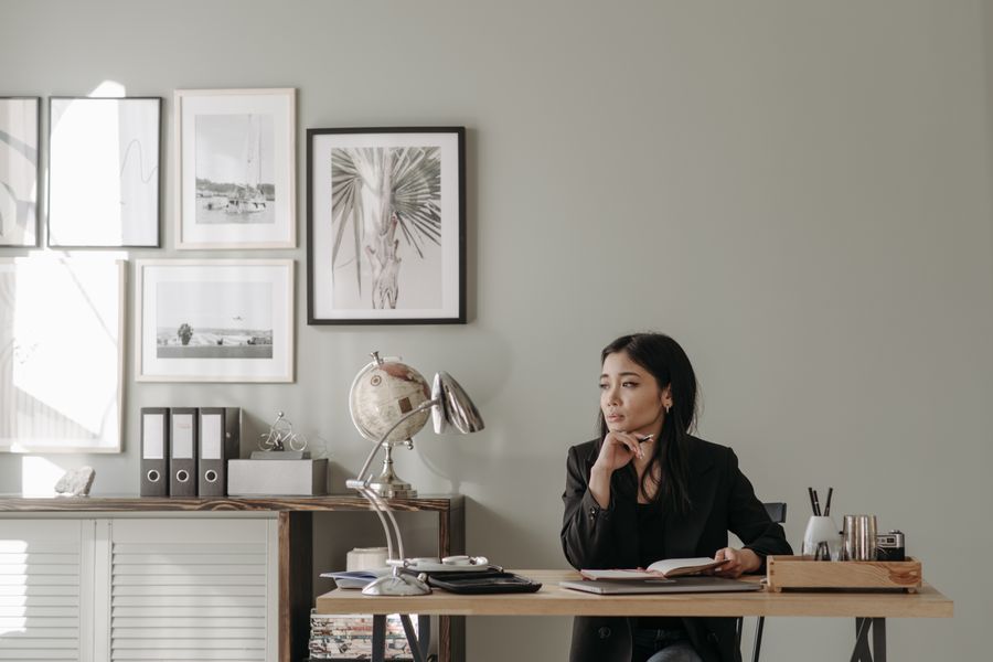 Asian woman in black blazer sitting at a desk in stylish home office, deep in thought.
