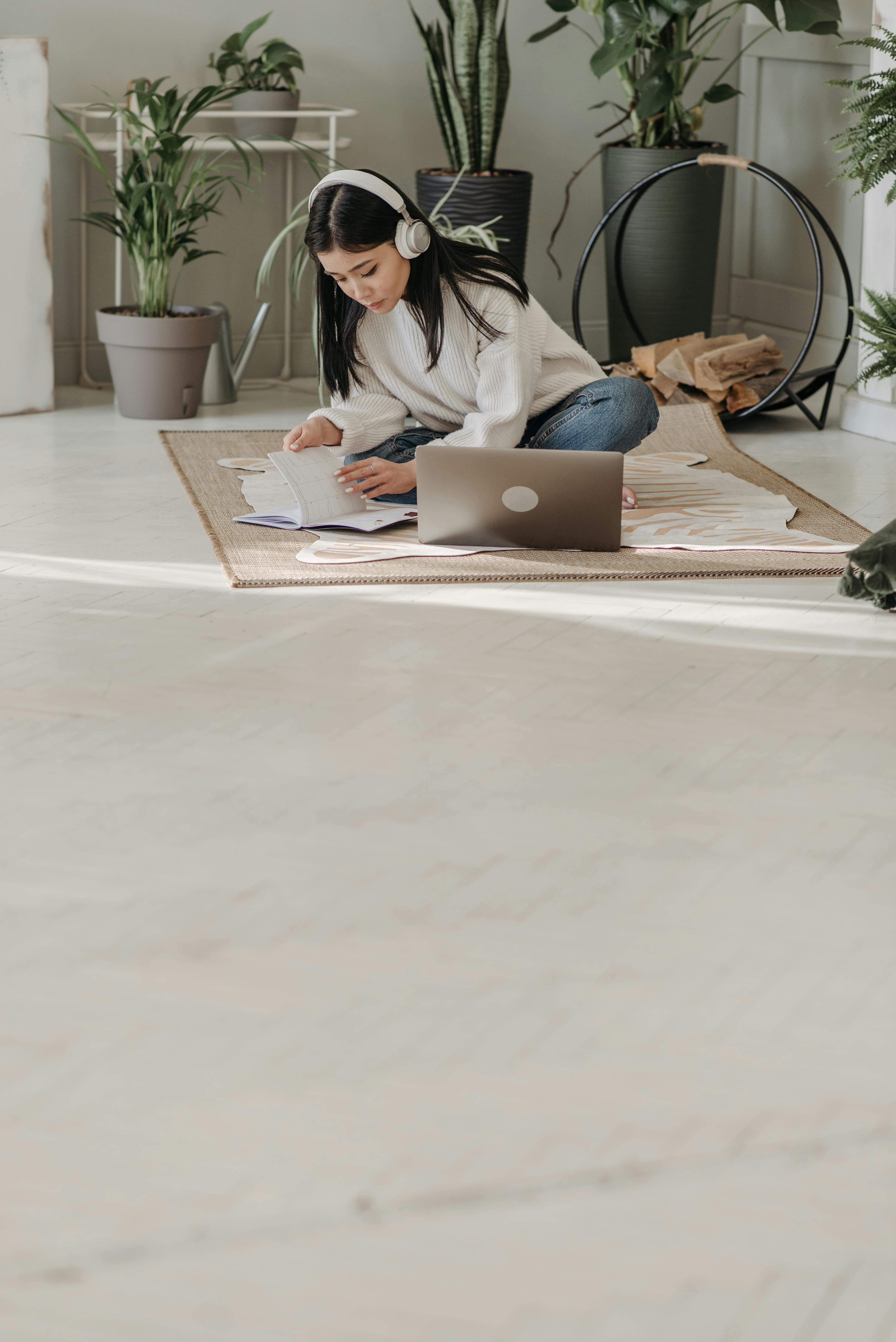 Free Young woman with headphones studies on floor with laptop, surrounded by plants, enjoying natural light. Stock Photo