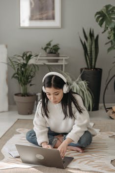 Focused young woman using laptop with headphones, sitting on floor, working remotely.