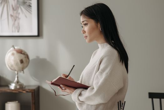 Close-up of a thoughtful woman writing in a notebook, wearing a knitted sweater indoors.