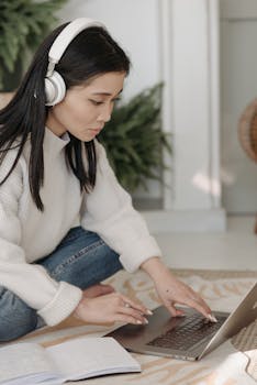 A woman wearing headphones works on her laptop at home, focused and diligent.