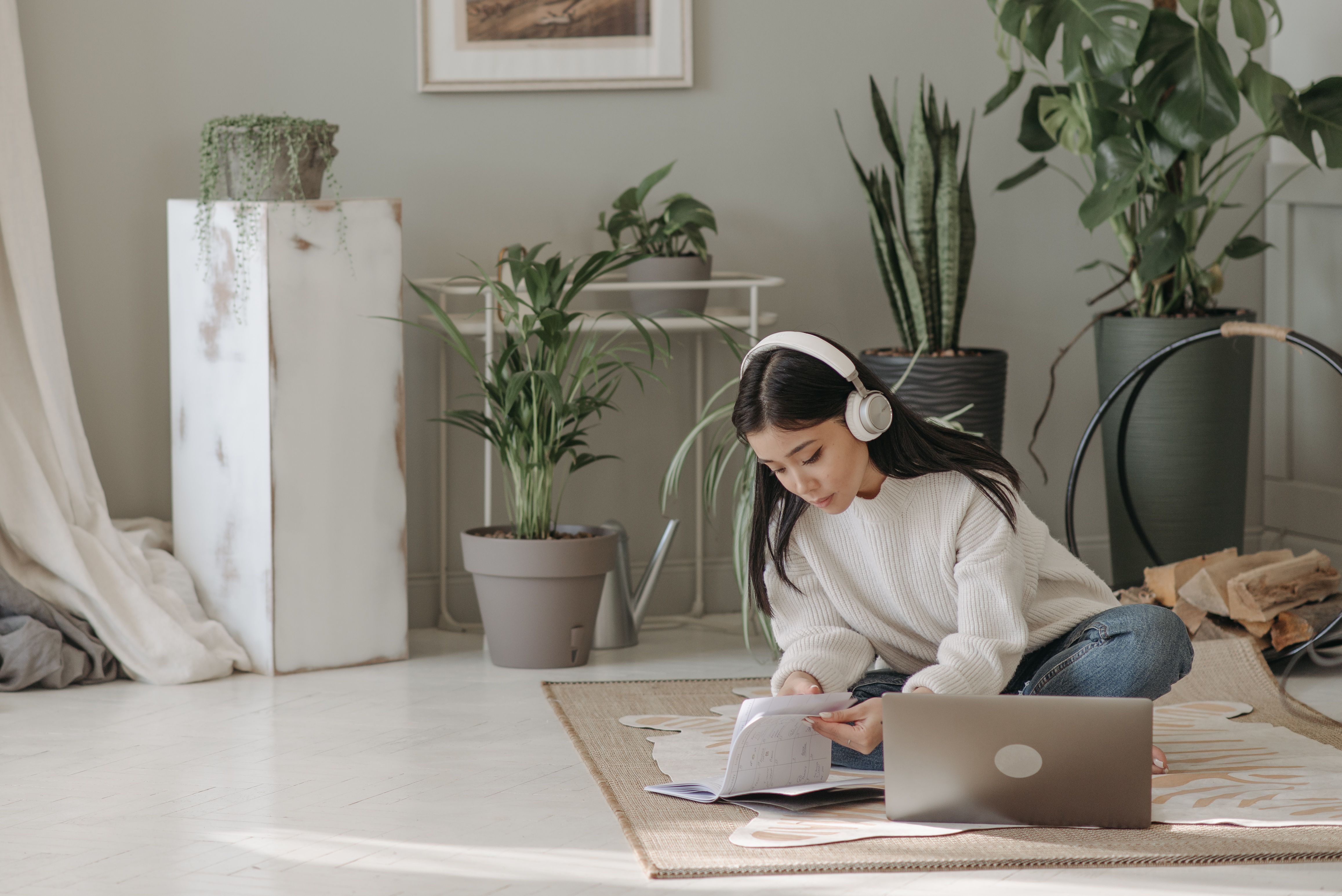 Asian woman seated on floor, using laptop and headphones, surrounded by indoor plants.